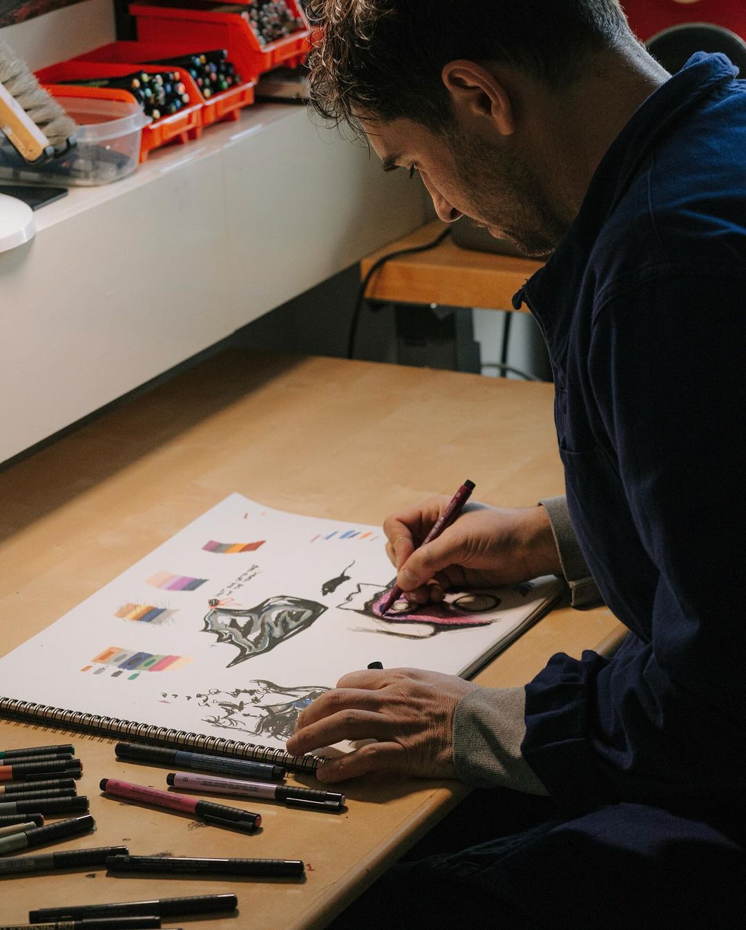 An artist sketches on a drawing pad with markers on a desk, wearing a blue shirt, creating detailed illustrations with precision in a workspace.