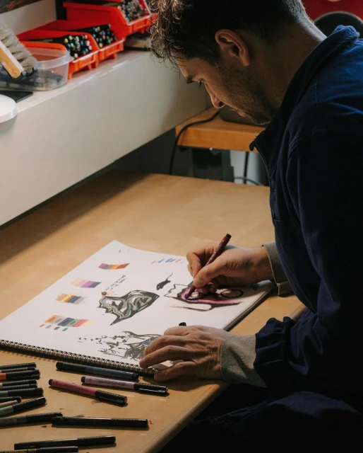 An artist sketches on a drawing pad with markers on a desk, wearing a blue shirt, creating detailed illustrations with precision in a workspace.