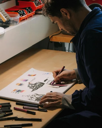 An artist sketches on a drawing pad with markers on a desk, wearing a blue shirt, creating detailed illustrations with precision in a workspace.
