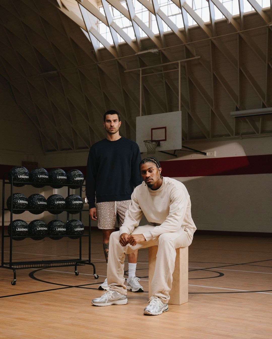Two adult males in a basketball court. One is sitting on a wooden block in a cream sweat suit, while the other stands behind him.
