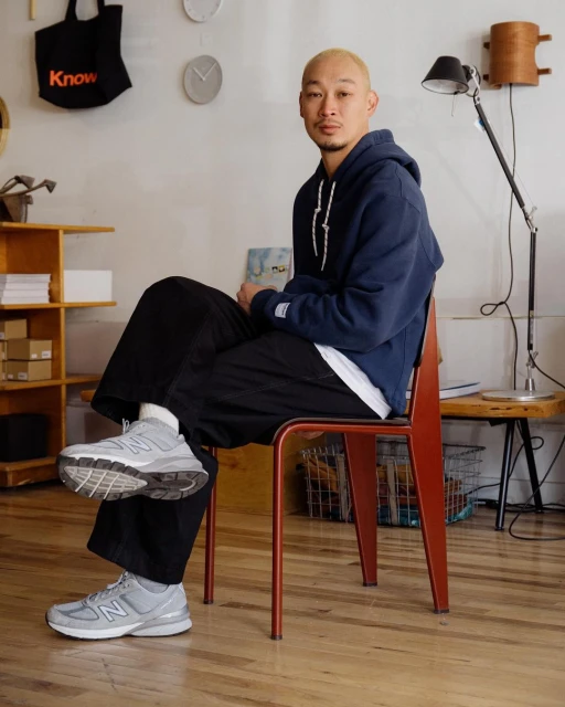 Adult male in hoodie and trousers sitting on a chair in an indoor setting with wood flooring. Light gray New Balance sneakers are visible.