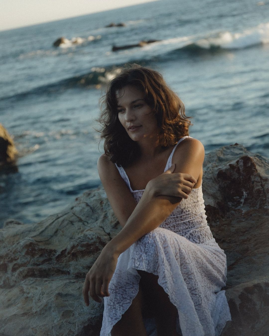 A brunette woman in a white dress sitting on rocks by the ocean, enjoying a serene moment with waves gently lapping in the background.