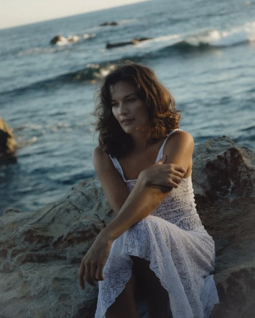 A brunette woman in a white dress sitting on rocks by the ocean, enjoying a serene moment with waves gently lapping in the background.