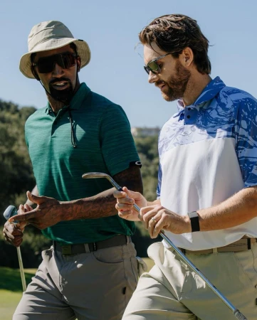 Two men wearing polo shirts and sunglasses on a golf course, holding golf clubs and smiling in sunny weather.