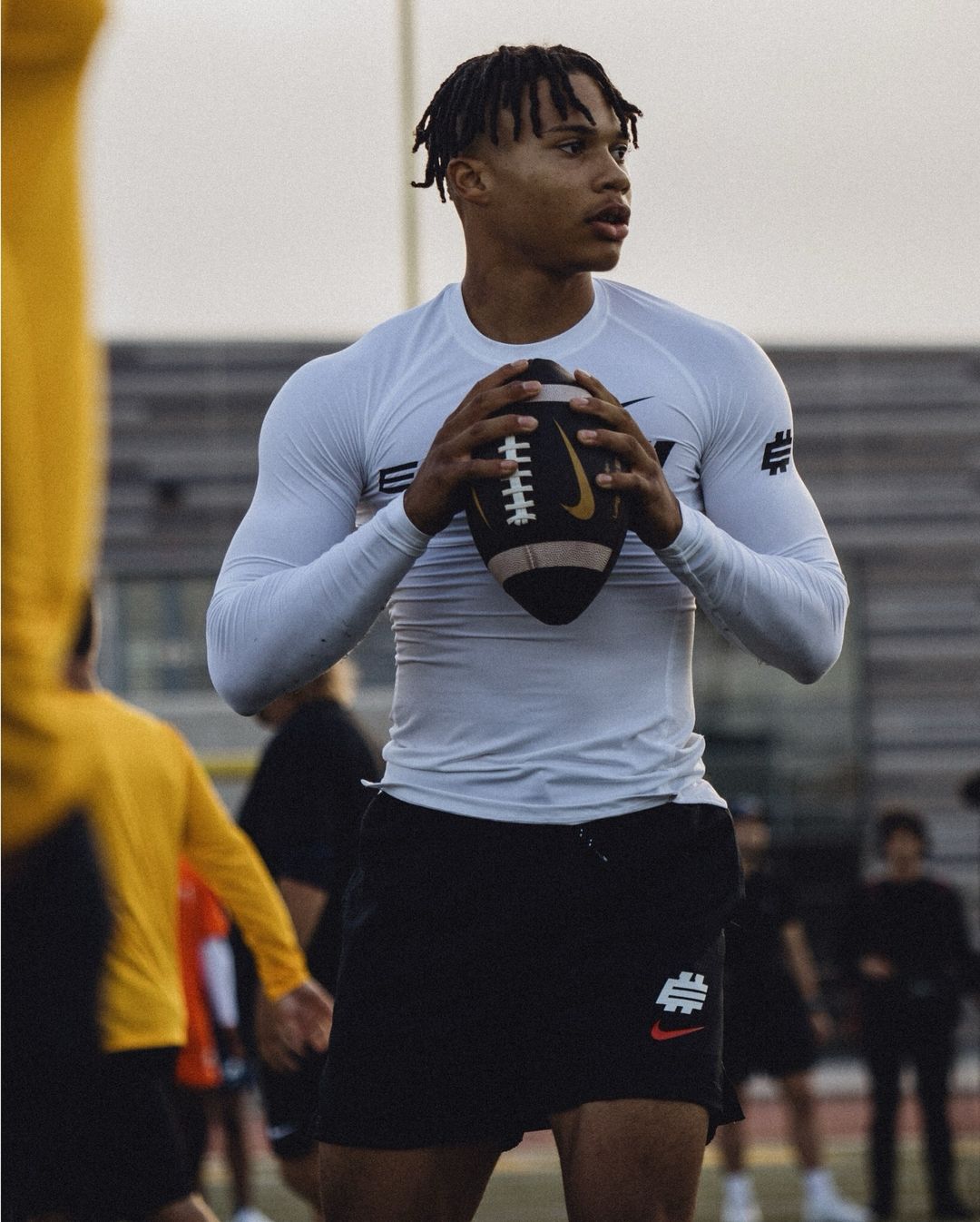 A muscular football player in athletic wear holds a football on a field, captured at eye-level with a shallow depth of field.