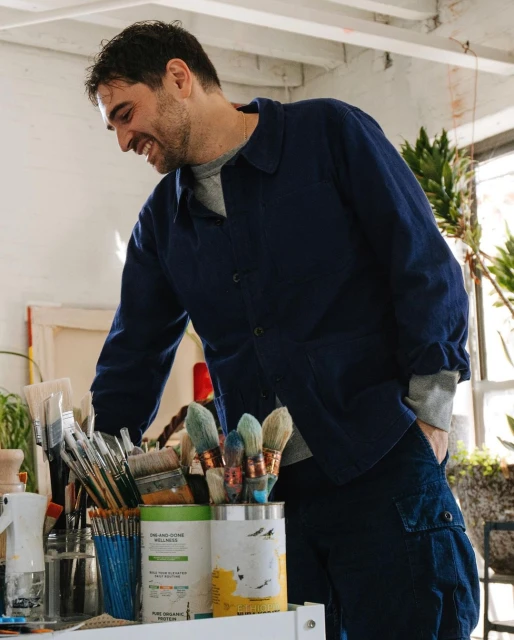 Smiling artist stands near an array of paintbrushes and supplies in a bright, airy studio setting, wearing a blue jacket and trousers.