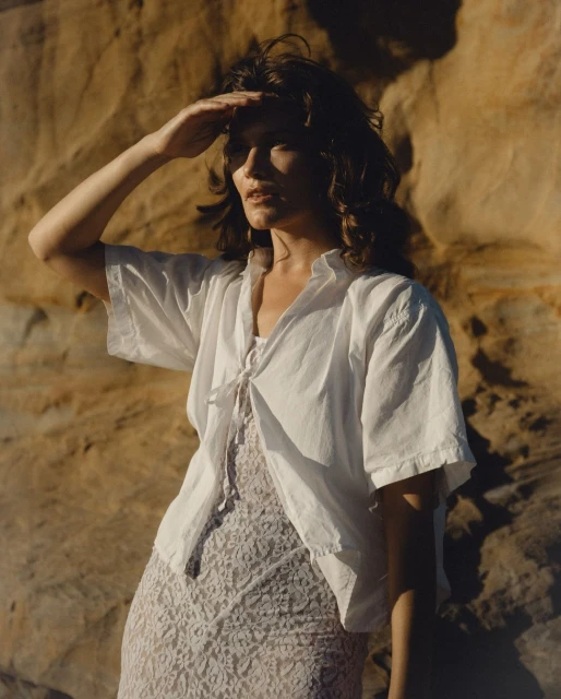 Woman in white blouse shields eyes in warm sunlight. Sandy rock background, casual style, looking into distance.