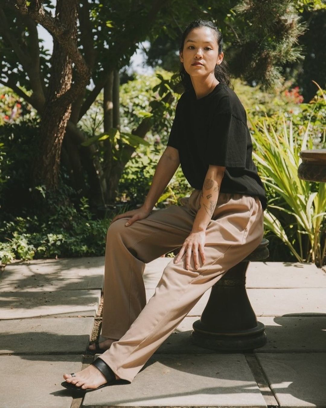 A woman sits on a pedestal in a garden wearing a black t-shirt and tan pants. The natural light highlights her features.