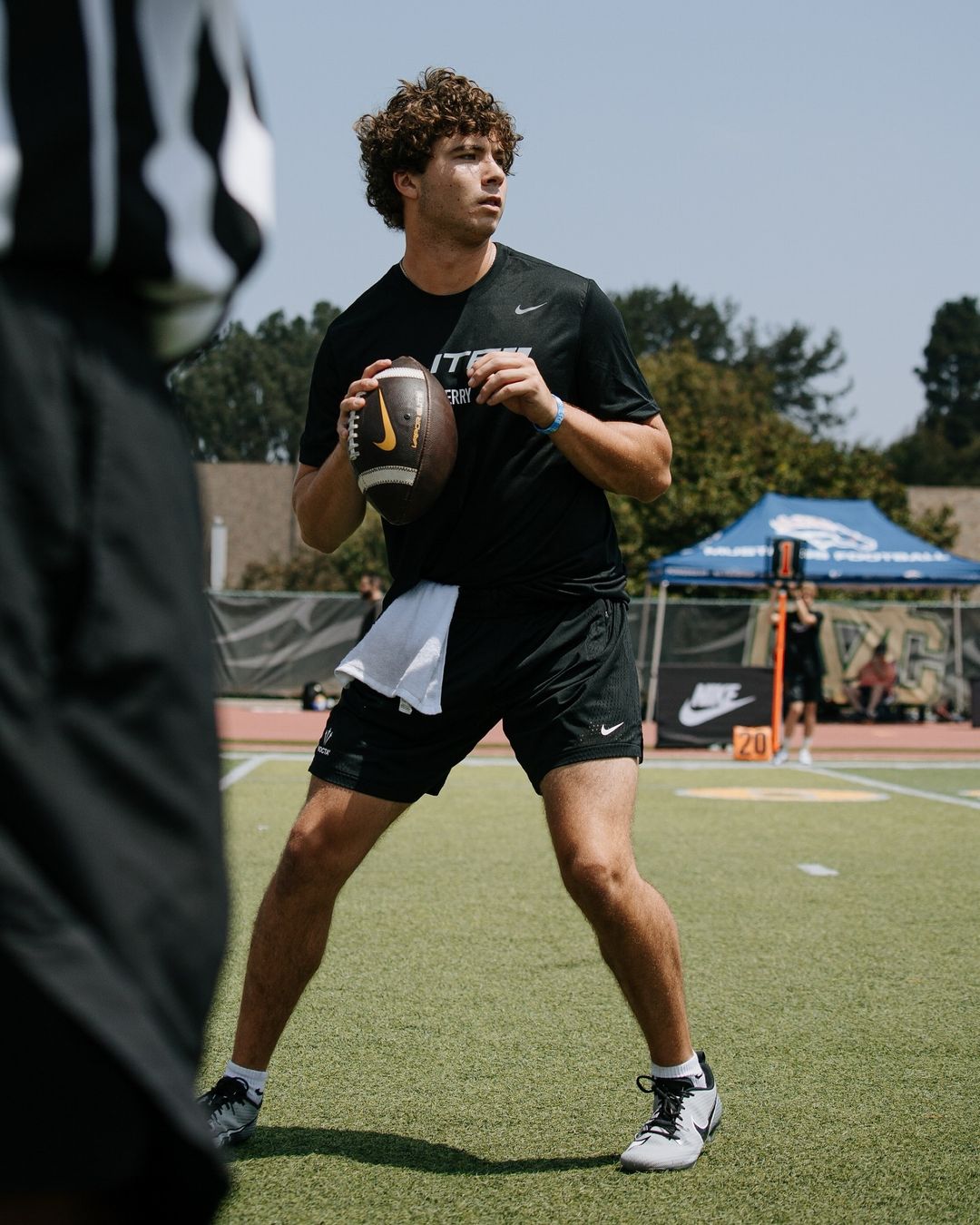 A male football player in a black uniform throws a football outdoors on a sunny day. The background features tents and greenery.