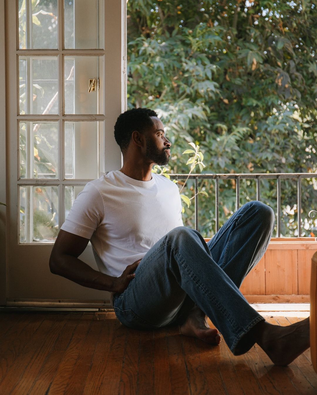 Indoor portrait of African-American man sitting on hardwood floor looking out window in white t-shirt and blue jeans enjoying the day.