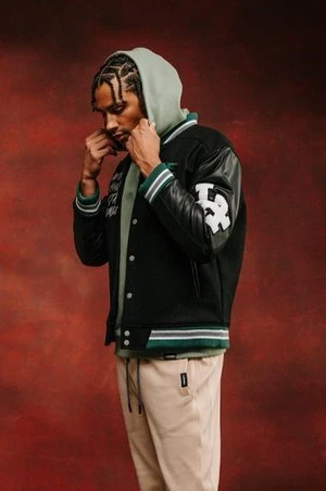 A man in a varsity jacket and hoodie strikes a pose against a textured backdrop. He wears tan pants with a serious expression.