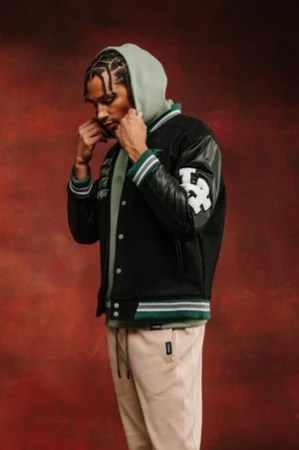 A man in a varsity jacket and hoodie strikes a pose against a textured backdrop. He wears tan pants with a serious expression.