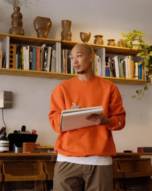 Man in orange sweatshirt holds a notebook and pen in an indoor space with wooden desk, bookshelf, and plants. Natural light.