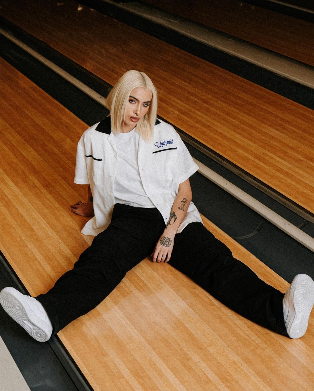 Woman with blond hair poses seated in white shirt, black pants and sneakers against vintage bowling alley background for fashion shoot.