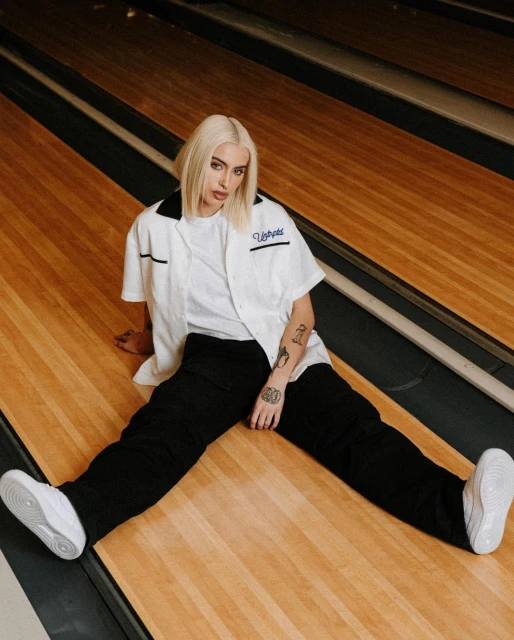 Woman with blond hair poses seated in white shirt, black pants and sneakers against vintage bowling alley background for fashion shoot.