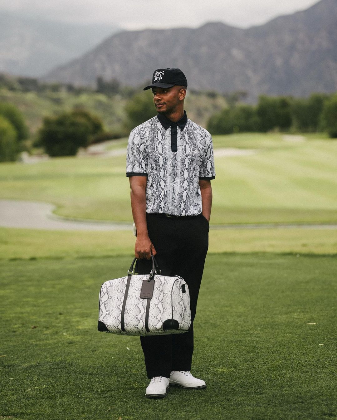 A man in a snakeskin polo shirt holds a matching duffel bag on a golf course with rolling green hills and mountains in the background.