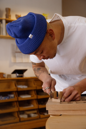 An adult working indoors uses a chisel and wood block on a woodworking project, wearing a blue knit cap and white shirt.