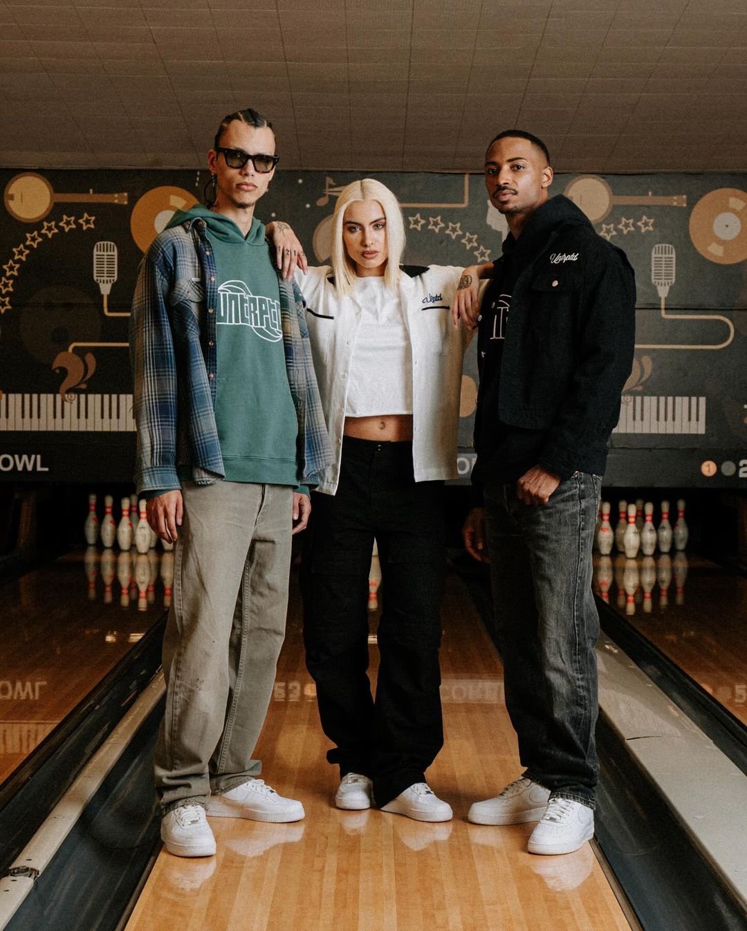 Three young adults in casual streetwear posing indoors at a bowling alley, showcasing contemporary fashion and lifestyle in a retro setting.