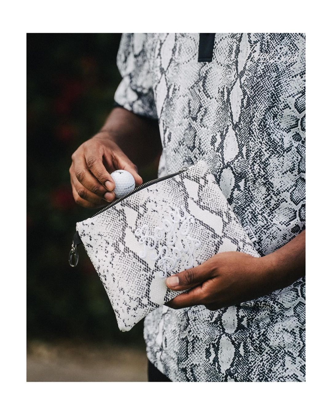 Person holds a golf ball over a snakeskin-patterned pouch outside. Snakeskin pattern shirt is visible. Dark blurred background.