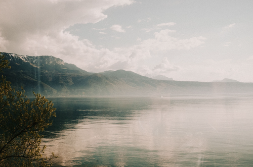 Scenic view of a calm lake reflecting mountains under a cloudy sky, framed by foliage in the foreground, creating a serene landscape.