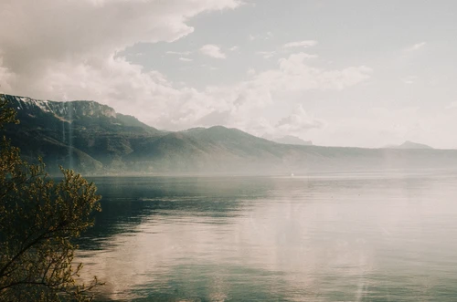 Scenic view of a calm lake reflecting mountains under a cloudy sky, framed by foliage in the foreground, creating a serene landscape.