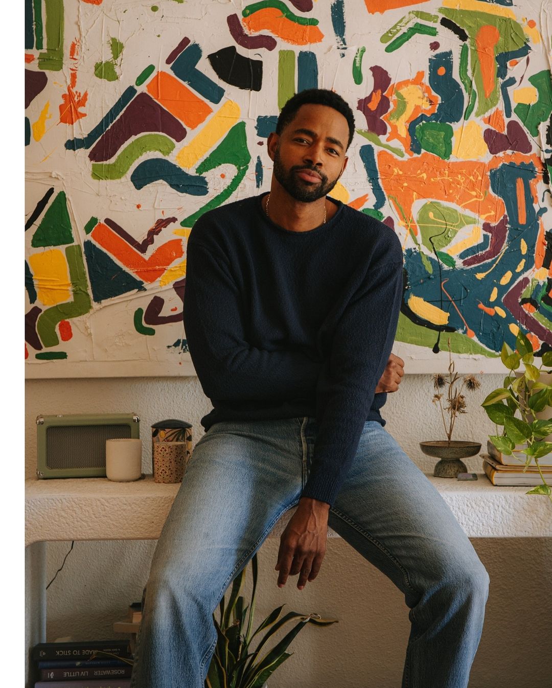 Portrait of an adult male sitting in an indoor setting, wearing jeans and a long-sleeved sweater, with a colorful painting in the background.