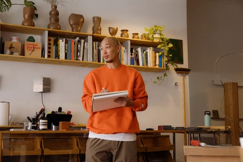 Adult male in an orange sweater stands in an artist studio, holding a notebook, with shelves of books and pottery in the background.