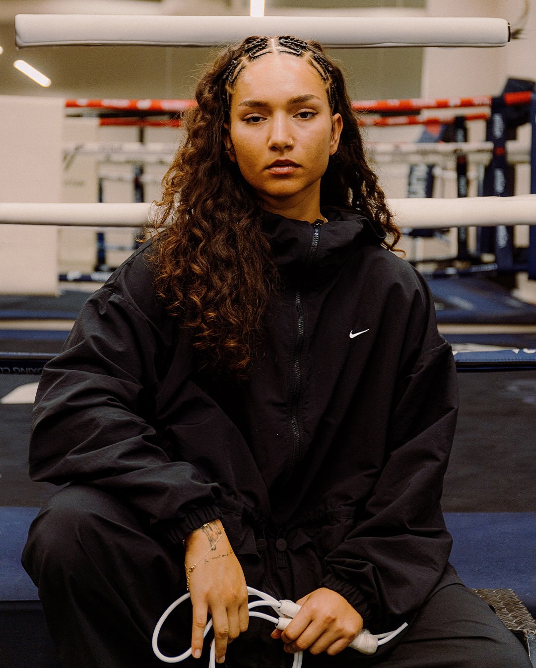 Female boxer in black athletic wear sits in boxing ring holding a jump rope, looking forward with a serious expression, indoor lighting.