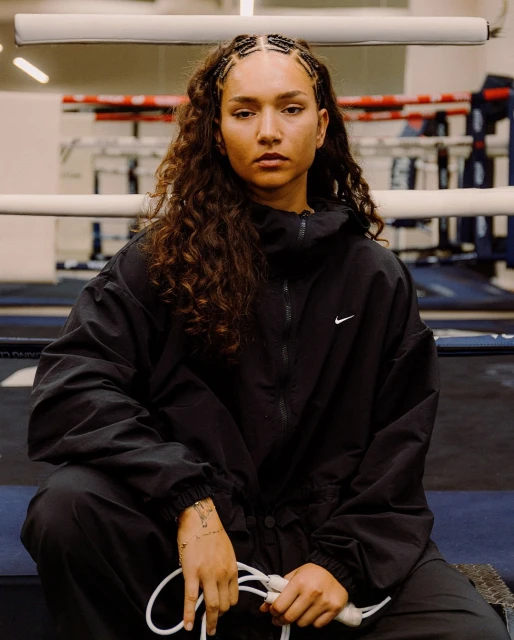 Female boxer in black athletic wear sits in boxing ring holding a jump rope, looking forward with a serious expression, indoor lighting.