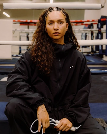 Female boxer in black athletic wear sits in boxing ring holding a jump rope, looking forward with a serious expression, indoor lighting.
