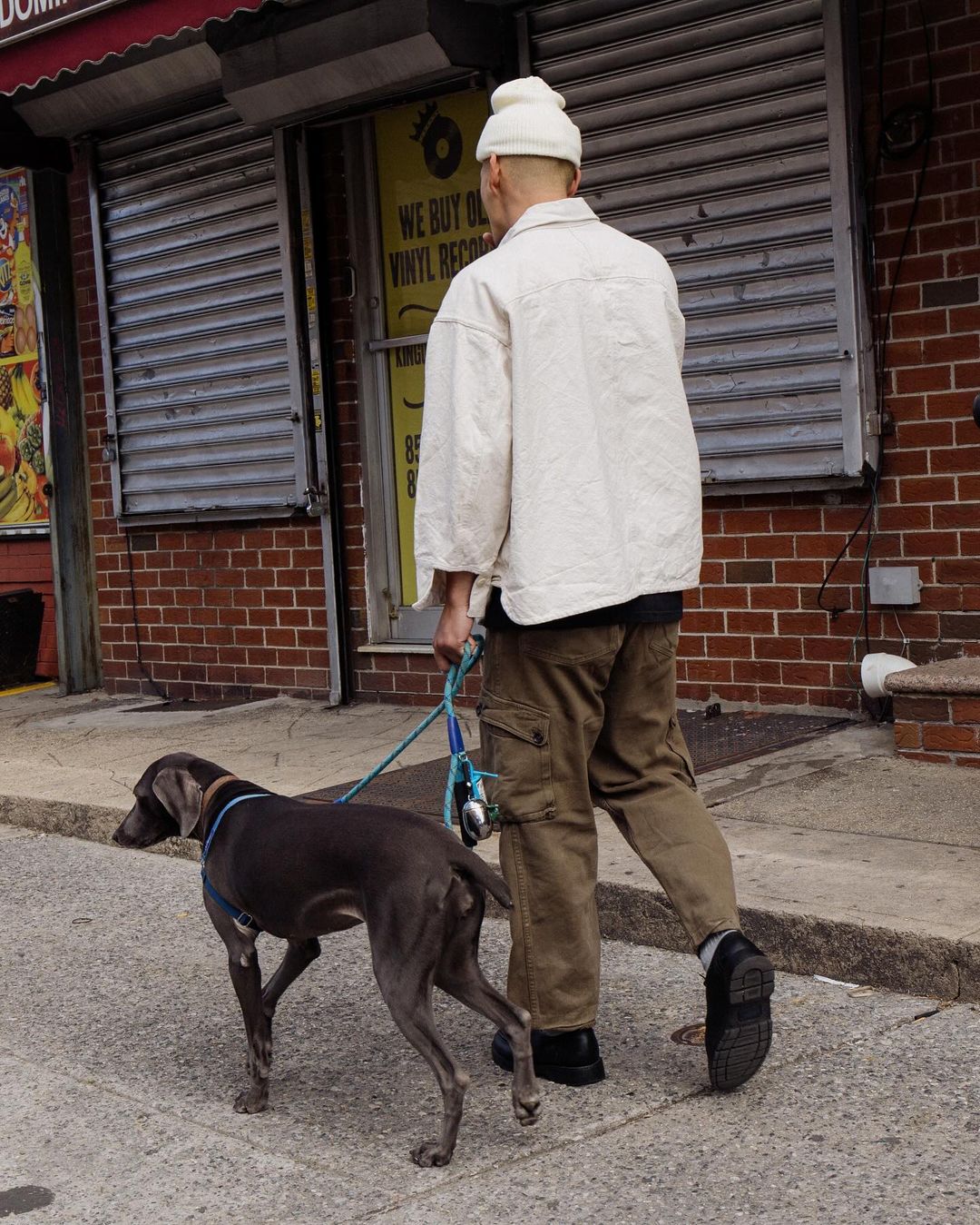 An adult walks a grey Weimaraner dog on a leash down a sidewalk next to a brick building with a closed, shuttered storefront.