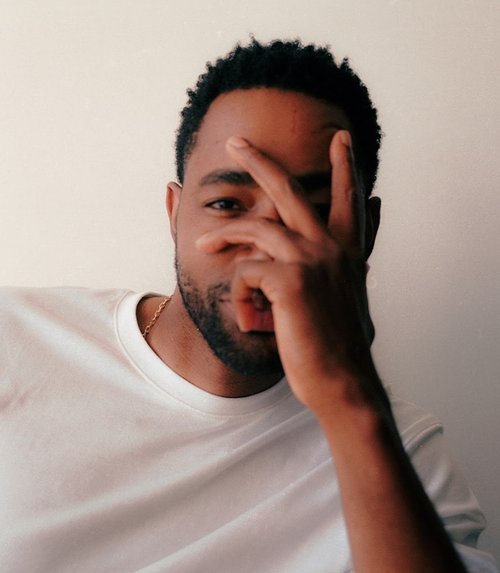 Portrait of an adult Black male wearing a white t-shirt, looking at the camera with his hand partially covering his face.