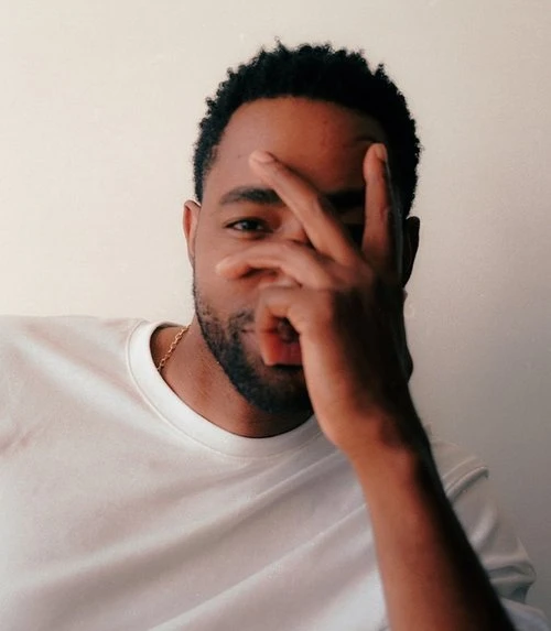 Portrait of an adult Black male wearing a white t-shirt, looking at the camera with his hand partially covering his face.