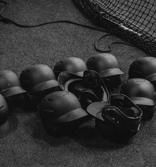 Pile of black baseball helmets, some upside down, on a textured gray surface, with a blurred black netting in the background.