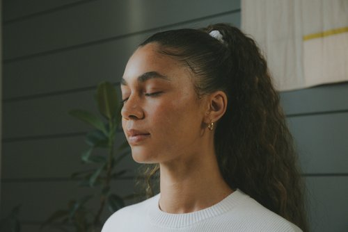 Woman with brown curly hair and a white sweater meditating with eyes closed and a plant in the background.