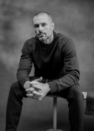 Black and white portrait of a man seated on a bench with hands clasped, wearing a dark crewneck sweatshirt and trousers against a mottled backdrop.