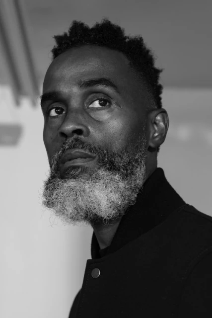 Black and white portrait of an African American man with a grey beard looking upward, wearing a black jacket against a white wall.