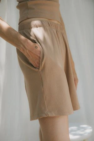 Close-up shot of a woman in a ribbed, light brown two-piece set, with her hand in the shorts pocket, against a soft white backdrop.