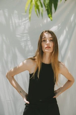 Woman in black top posing against a white draped backdrop with tropical leaf shadows above