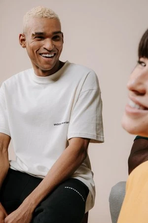 A light-skinned adult male with blond hair smiles while wearing a white tee shirt and black pants, in a close-up indoor shot.