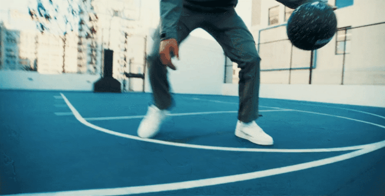 A person dribbles a basketball on an outdoor blue court surrounded by buildings on a sunny day, showcasing sport and urban activity.