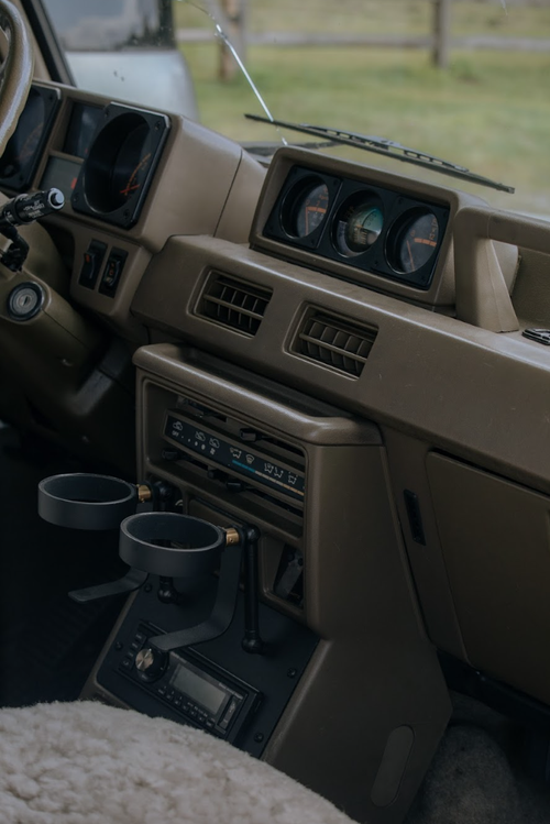 Automotive interior featuring a beige car dashboard with gauges, air vents, an audio system, cup holders, and a sheepskin seat cover.