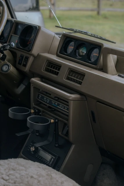 Automotive interior featuring a beige car dashboard with gauges, air vents, an audio system, cup holders, and a sheepskin seat cover.