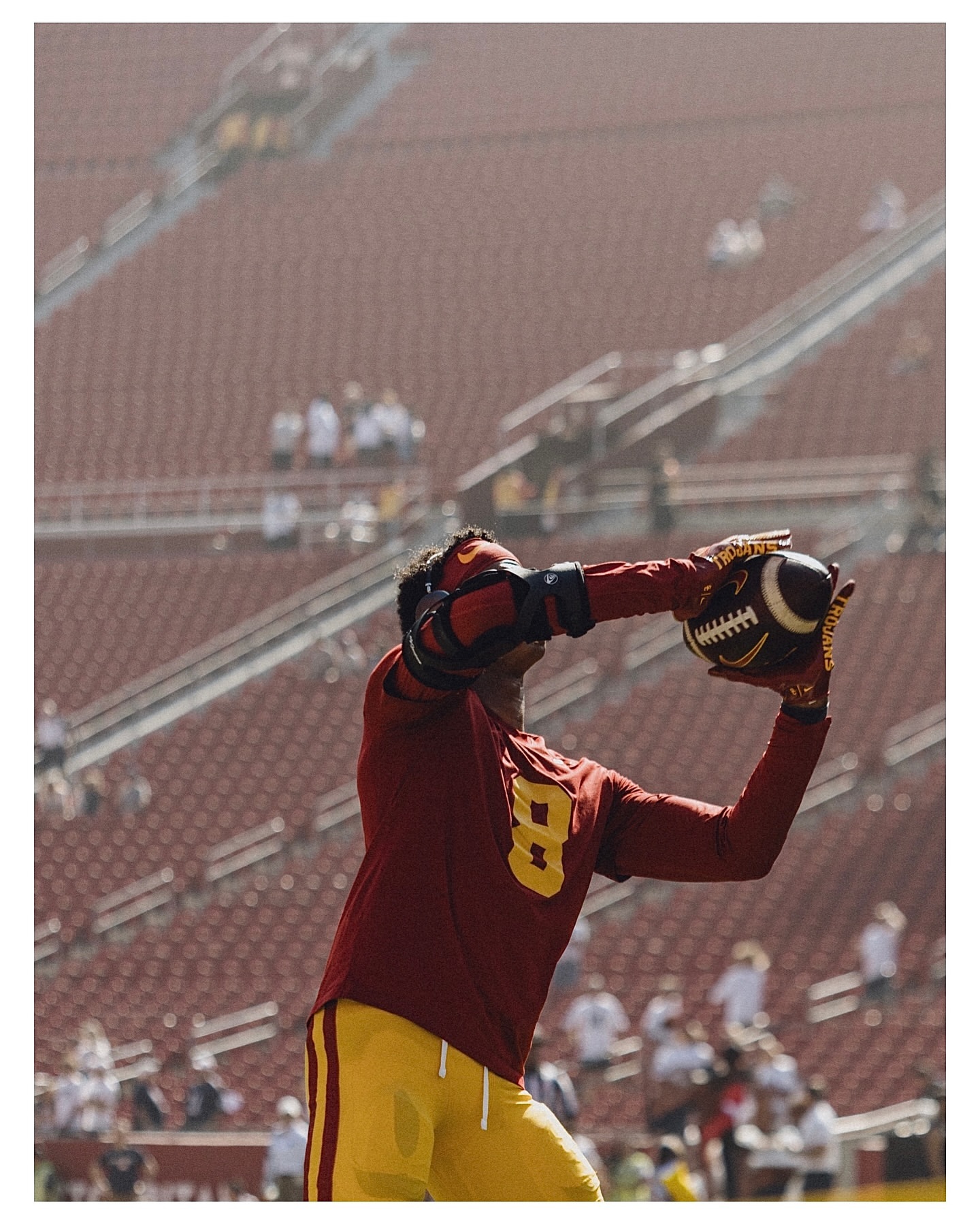 Football player in maroon and gold uniform reaches to catch a football during a game in a sunlit outdoor stadium.