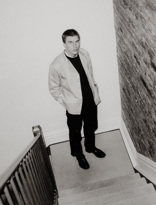 Monochrome portrait of a man standing on a staircase, wearing a black sweater and light-colored jacket, shot from a high angle.