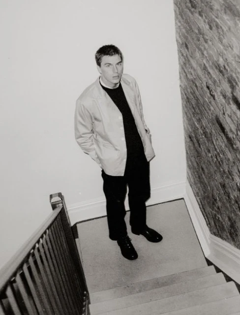 Monochrome portrait of a man standing on a staircase, wearing a black sweater and light-colored jacket, shot from a high angle.