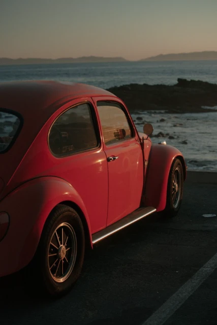 Red Volkswagen Beetle parked near a rocky coastline at sunset, capturing a vintage car against a scenic seascape during golden hour.