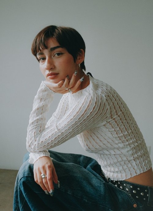 Woman with short dark hair wearing a white textured long-sleeve top and blue denim jeans against a plain wall