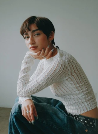 Woman with short dark hair wearing a white textured long-sleeve top and blue denim jeans against a plain wall