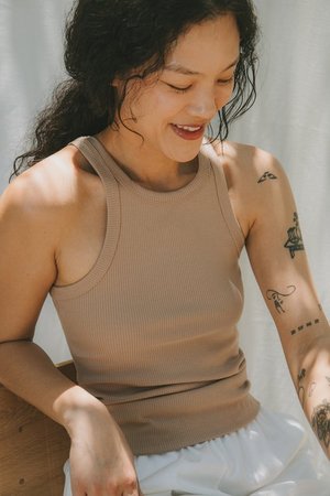 Smiling young Asian woman in ribbed beige tank top with arm tattoos, sitting indoors with natural light, looking downwards.